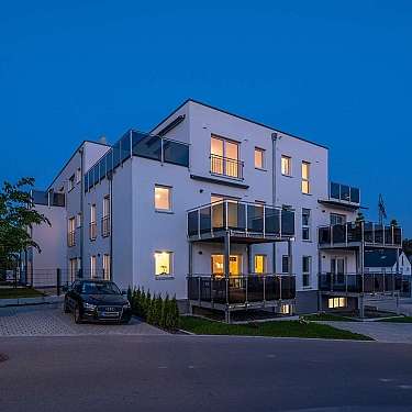 Modernes Mehrfamilienhaus mit beleuchteten Fenstern und Balkonen bei Dämmerung.