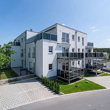 Modernes weißes Mehrfamilienhaus mit Glasbalkonen und gepflastertem Vorplatz.