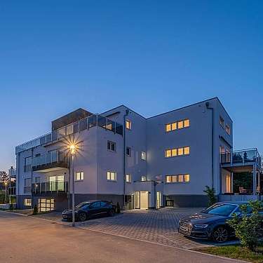 Modernes Mehrfamilienhaus mit beleuchteten Fenstern, Balkonen und geparkten Autos bei Dämmerung.
