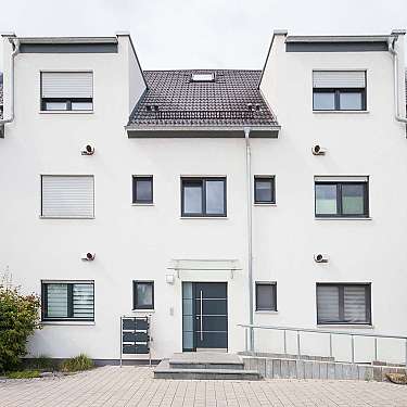 Weiße Hausfassade mit mehreren Fenstern, dunkel gedecktem Dach und Eingangsbereich.